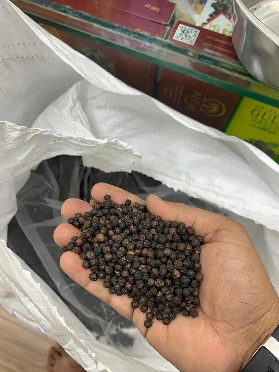 Black peppercorns held in an open hand above a sack of pepper